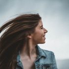 Portrait of a smiling young woman in a denim jacket against a cloudy sky.