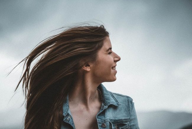 Portrait of a smiling young woman in a denim jacket against a cloudy sky.
