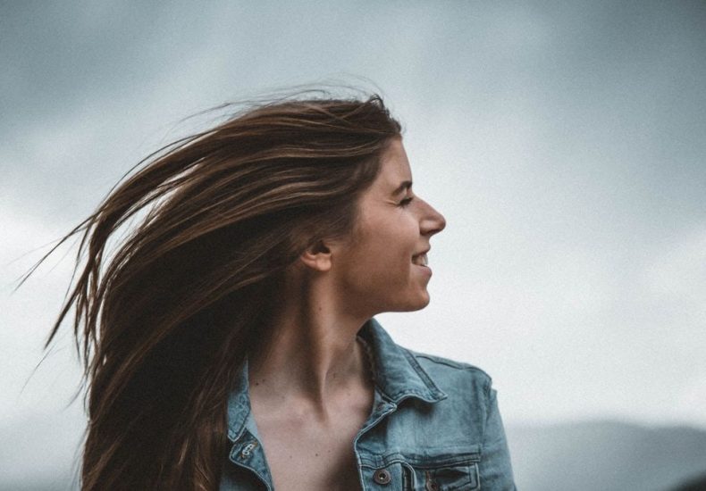 Portrait of a smiling young woman in a denim jacket against a cloudy sky.