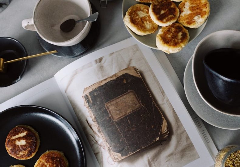 Top view of plate with delicious curd pancakes placed on recipe book surrounded by various plates on gray tabletop in kitchen