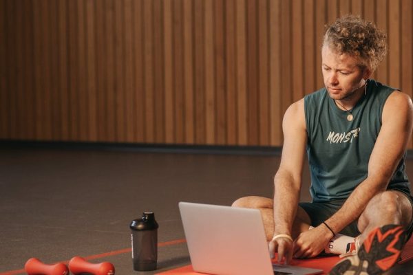 Man sitting on exercise mat with laptop, surrounded by dumbbells, creates a unique fitness workspace.