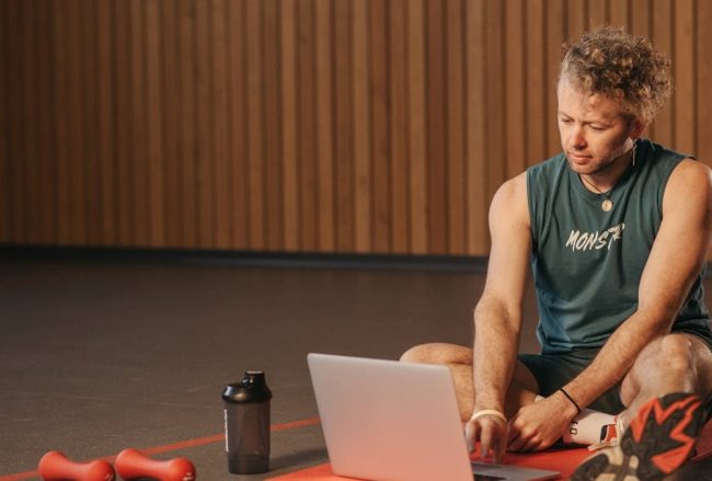 Man sitting on exercise mat with laptop, surrounded by dumbbells, creates a unique fitness workspace.